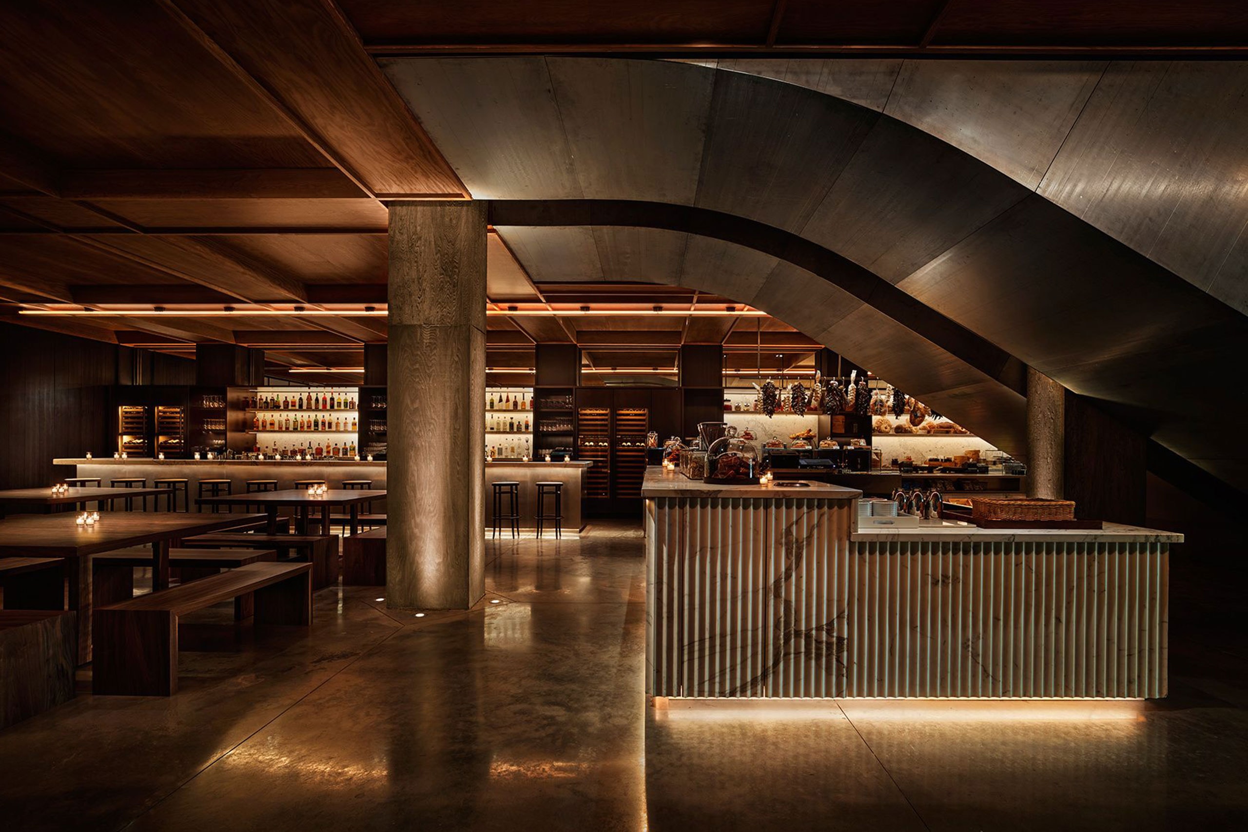 A sophisticated bar and dining space at PUBLIC Hotel New York, featuring SENTIENT’s custom-made communal tables and benches. The contemporary design includes dim ambient lighting, a sculptural staircase, and an open-concept bar with an extensive selection of spirits. The combination of raw materials, warm wood tones, and modern industrial elements creates a stylish and inviting atmosphere.