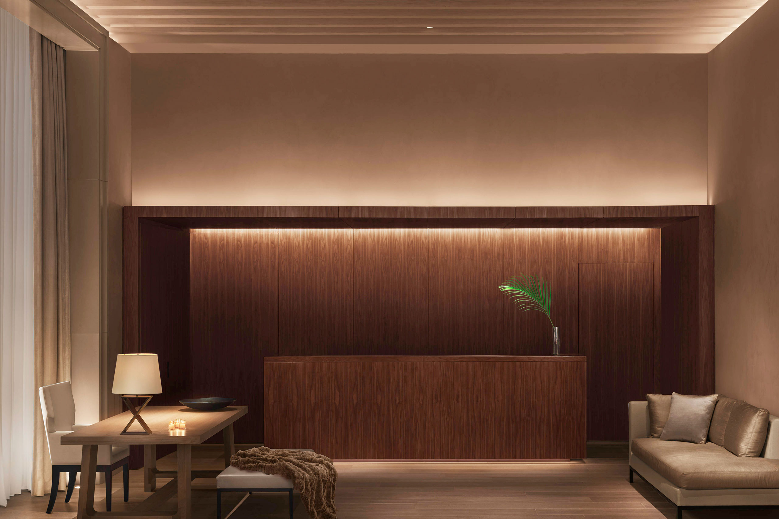 A serene and minimalist hotel lobby at The New York Edition, featuring custom furniture by SENTIENT. The design includes a sleek walnut reception desk with warm ambient lighting, a refined wooden table with a sculptural base, and a plush seating area. The soft neutral tones and modern aesthetic create a welcoming and sophisticated atmosphere.