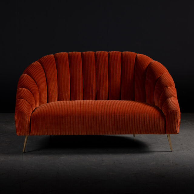 The SENTIENT Study Sofa upholstered in vibrant orange corduroy fabric, featuring a scalloped backrest and brass legs. A bold and luxurious seating piece for modern and eclectic interiors.