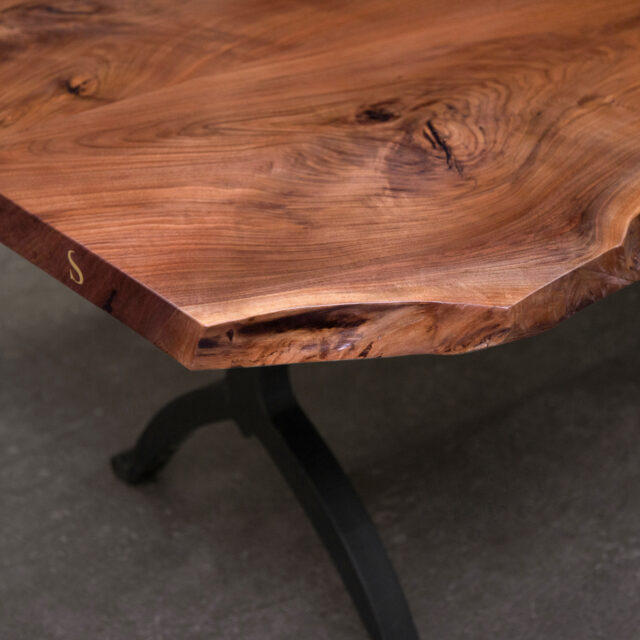 A close-up of a SENTIENT live edge walnut table featuring a natural wood grain top with a brass inlay detail. The table rests on a sturdy black metal base, blending rustic charm with modern elegance.