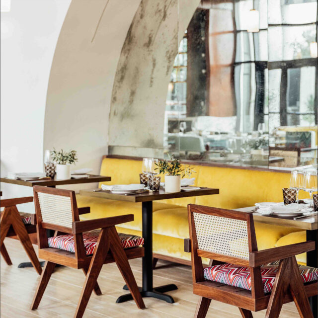 A vibrant restaurant interior featuring SENTIENT custom dining tables and wooden chairs with woven cane backs and patterned cushions. The design is complemented by a yellow banquette and an arched mirror, creating a stylish and inviting atmosphere.