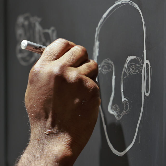 A close-up of an artist’s hand drawing a face on a chalkboard as part of the interactive exhibition featuring the Sapience Chair by SENTIENT.