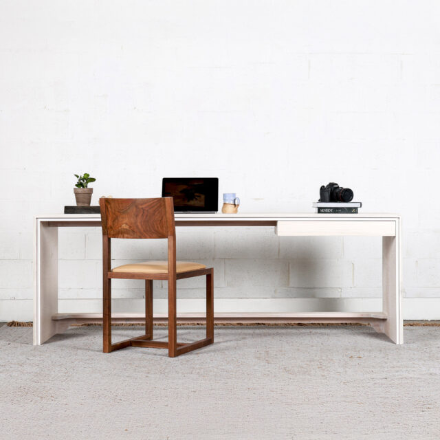 A minimally designed desk with a drawer agains a white wall with a walnut chair next to it. A laptop, a camera and a few books are on the desk