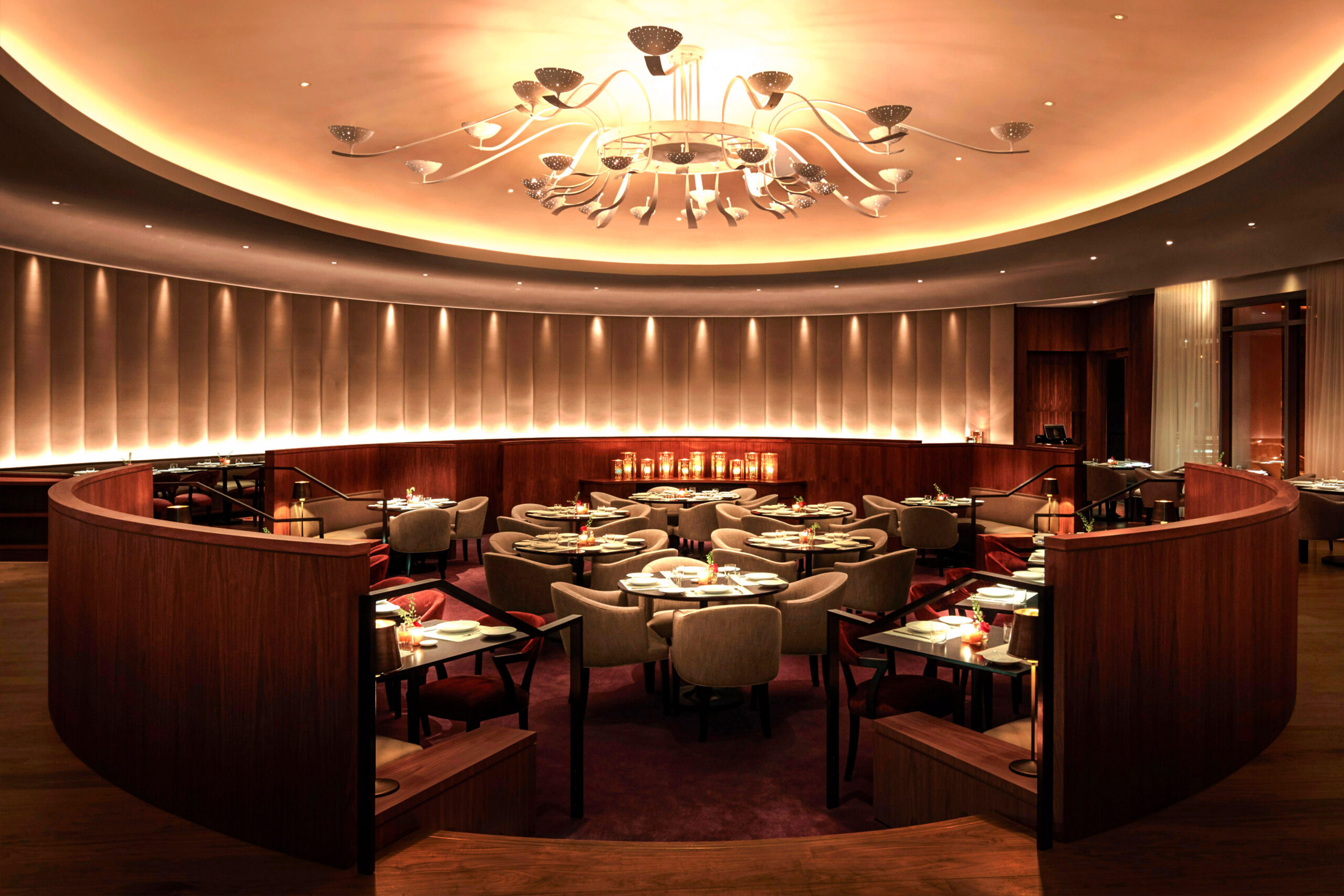 Custom Restaurant Furniture by SENTIENT – Hospitality Project Elegant restaurant interior featuring custom-made dining furniture by SENTIENT, with curved banquette seating, soft upholstered chairs, and minimalist black tables under ambient ceiling lighting and a sculptural chandelier.