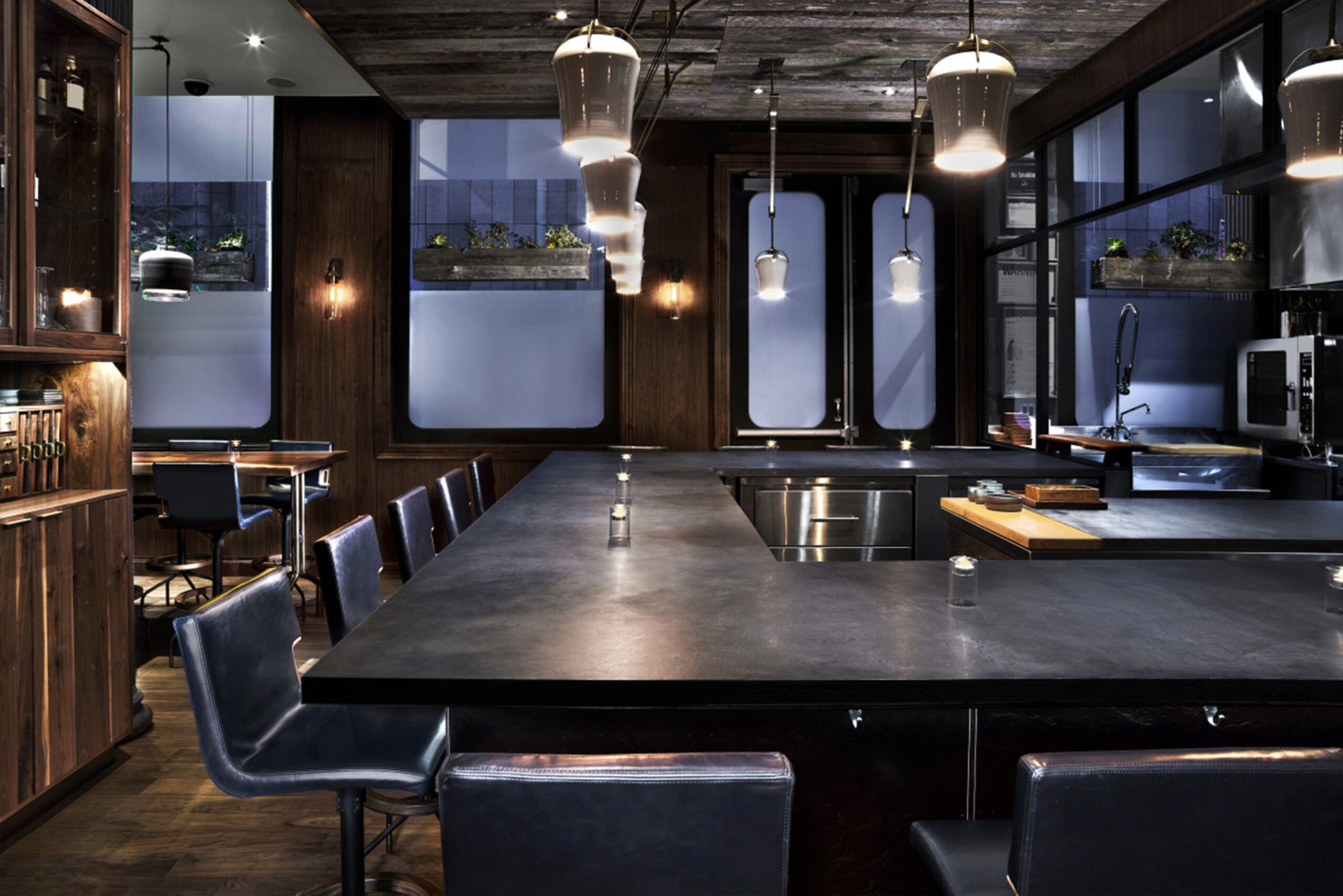 Custom Furnishings by SENTIENT for Atera Restaurant’s Open Kitchen Setting Custom-made furniture by SENTIENT at Atera Restaurant in New York, featuring a dark stone U-shaped chef’s counter surrounded by leather-upholstered bar stools in a moody, modern interior with wood paneling and pendant lighting.