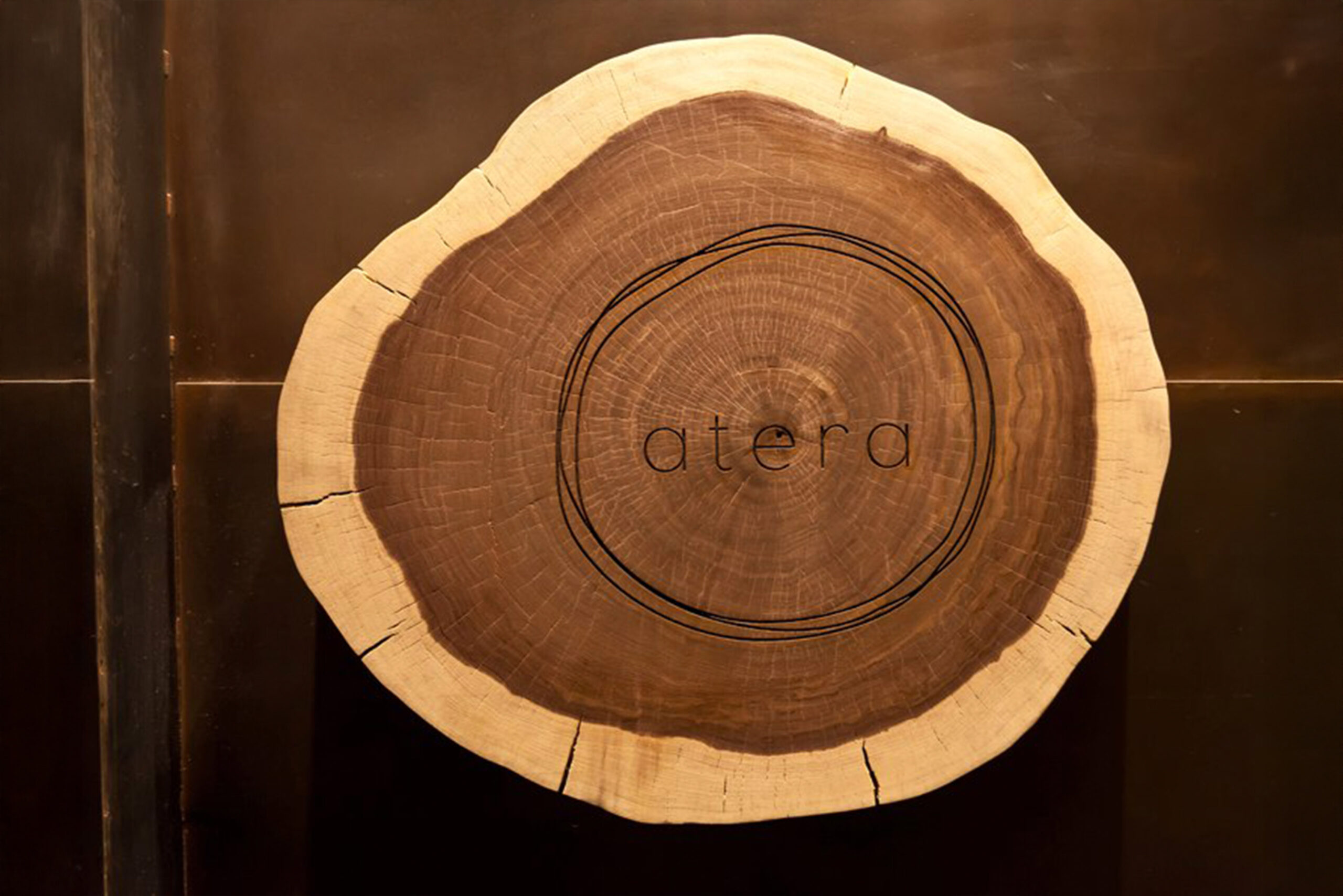 Atera Restaurant Sign Featuring Tree Cross-Section with Custom Branding Close-up of a natural wood tree cross-section with the word “atera” engraved in the center, showcasing custom branding and organic design elements at Atera Restaurant in New York.