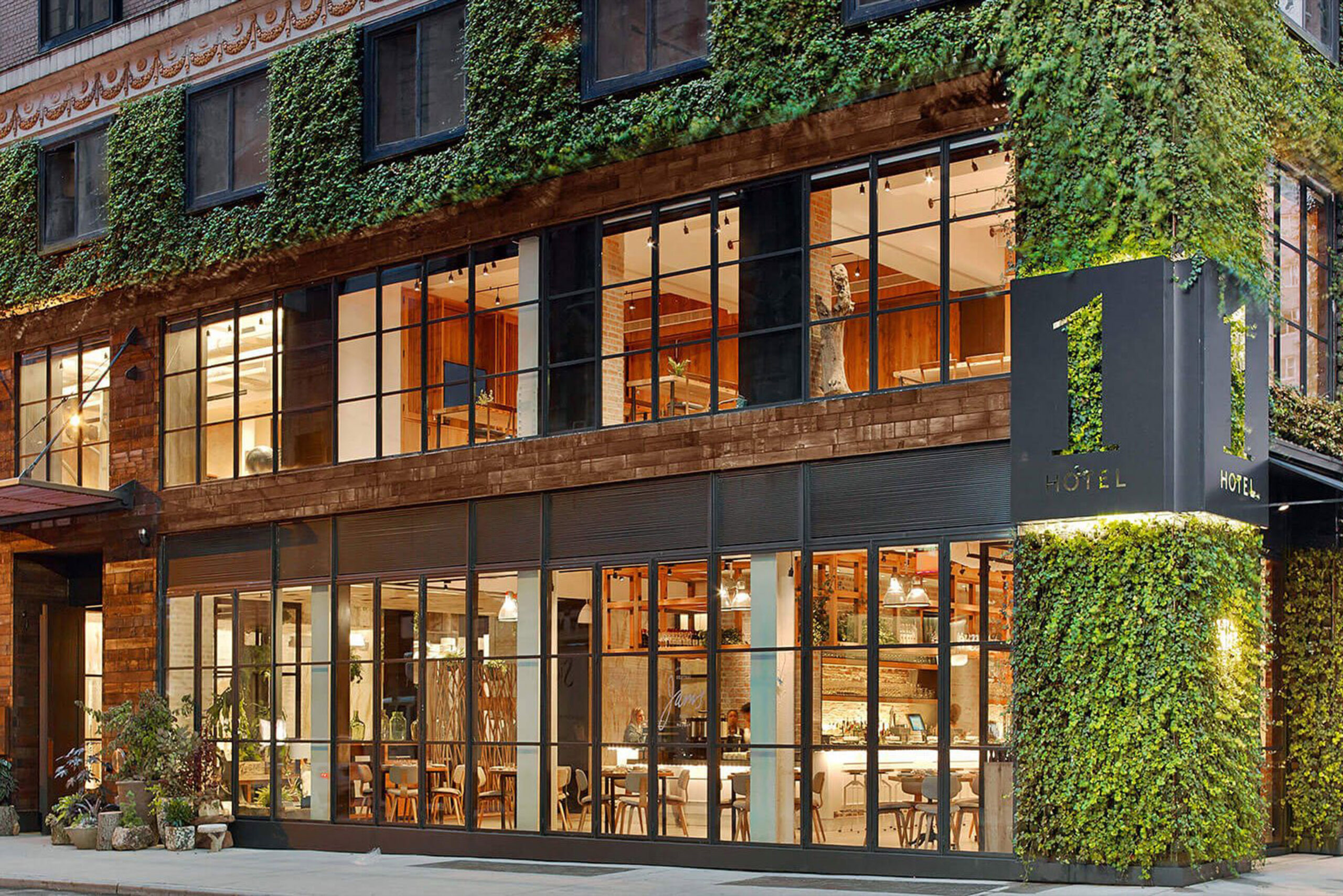 SENTIENT Custom Tables at Jams Restaurant, 1 Hotel Central Park NYC Exterior view of 1 Hotel Central Park in New York City, featuring the green living wall façade and warm interior of Jams Restaurant with custom-made wood tables by SENTIENT visible through the large windows.