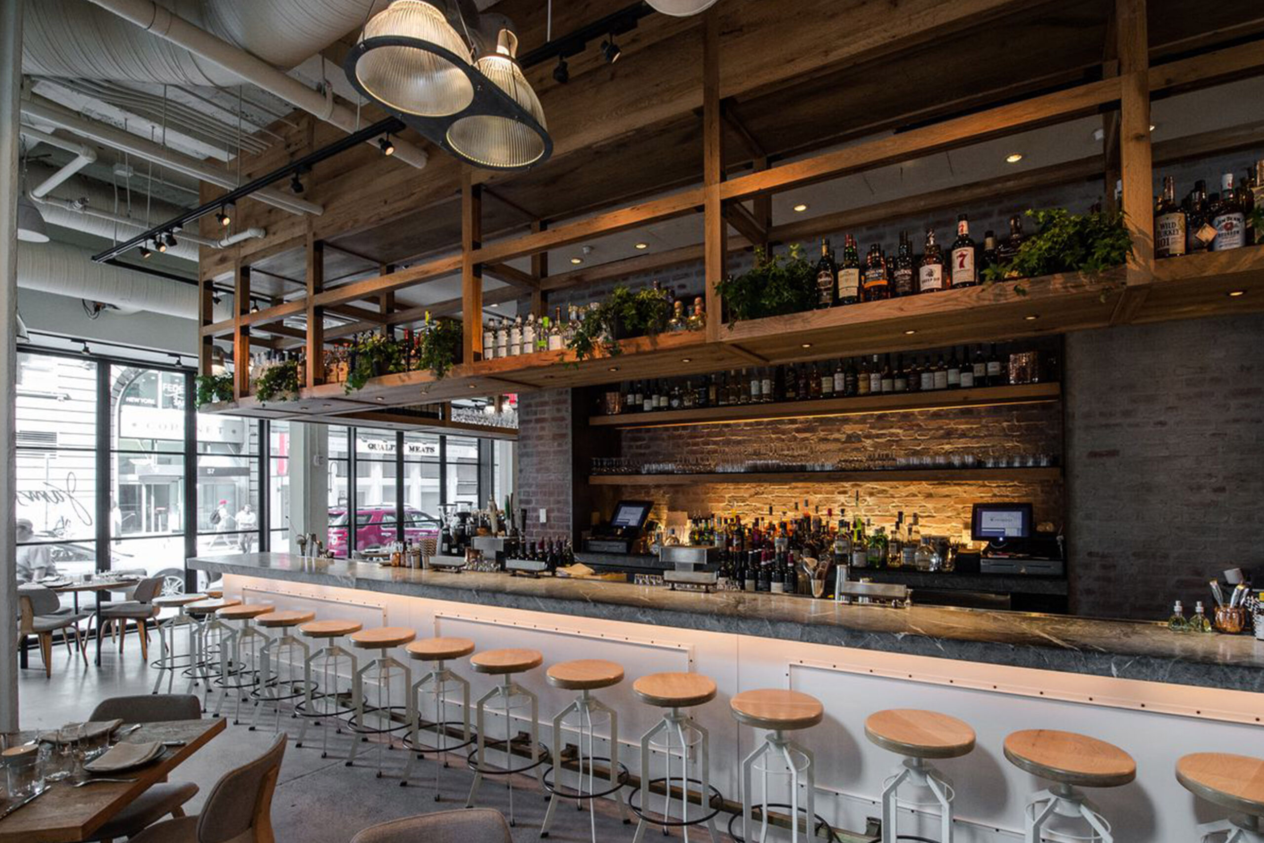 Custom Bar and Tables by SENTIENT – Jams Restaurant NYC Interior Modern bar interior at Jams restaurant in New York City featuring custom bar counter and tables by SENTIENT, with wood and metal bar stools, overhead shelving with greenery, and exposed brick and timber ceiling design.