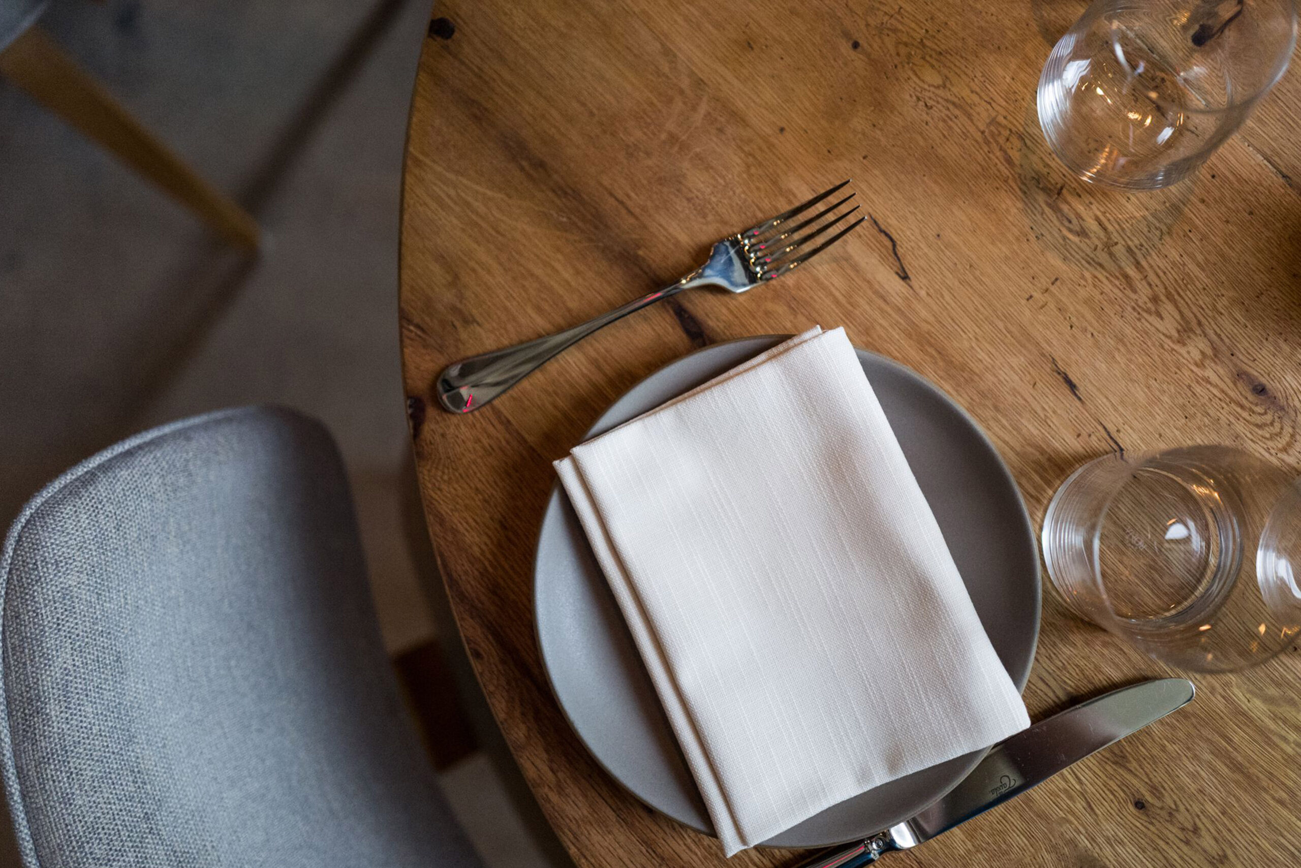Detail View – Custom Dining Table at Jams Restaurant by SENTIENT Close-up of custom round wood table at Jams restaurant in NYC by SENTIENT, featuring elegant table setting with neutral plate, white napkin, cutlery, and glassware on natural wood grain surface.