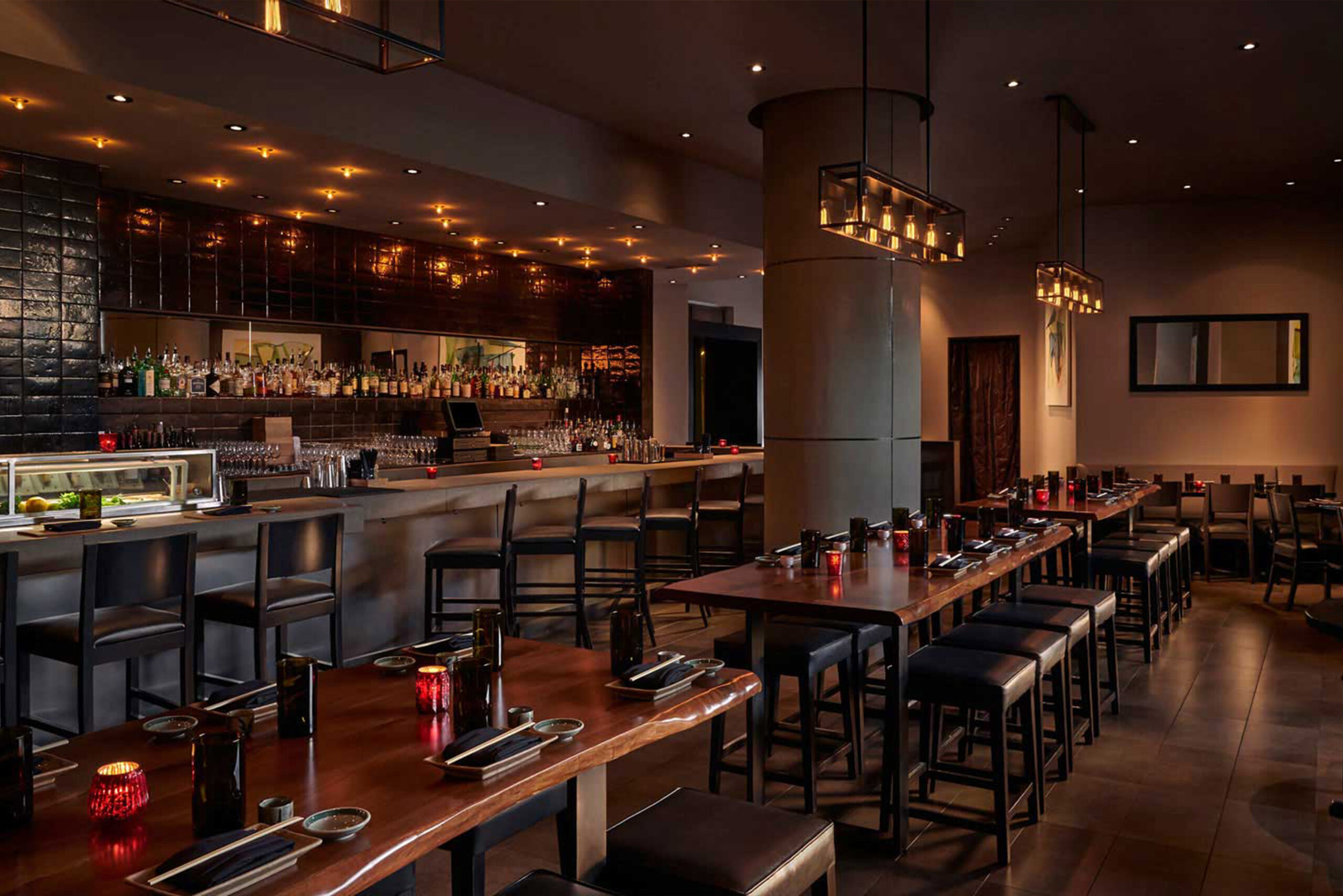 Custom Live Edge Tables by SENTIENT – Zentan at The Kimpton Donovan Hotel Elegant restaurant and bar interior at Zentan in The Kimpton Donovan Hotel, featuring custom live edge communal tables with dark wood stools by SENTIENT, moody lighting, and a fully stocked bar with black tile backdrop.
